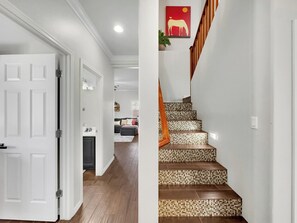 Un couloir avec une porte blanche, un escalier aux marches décorées et, au fond, un salon.