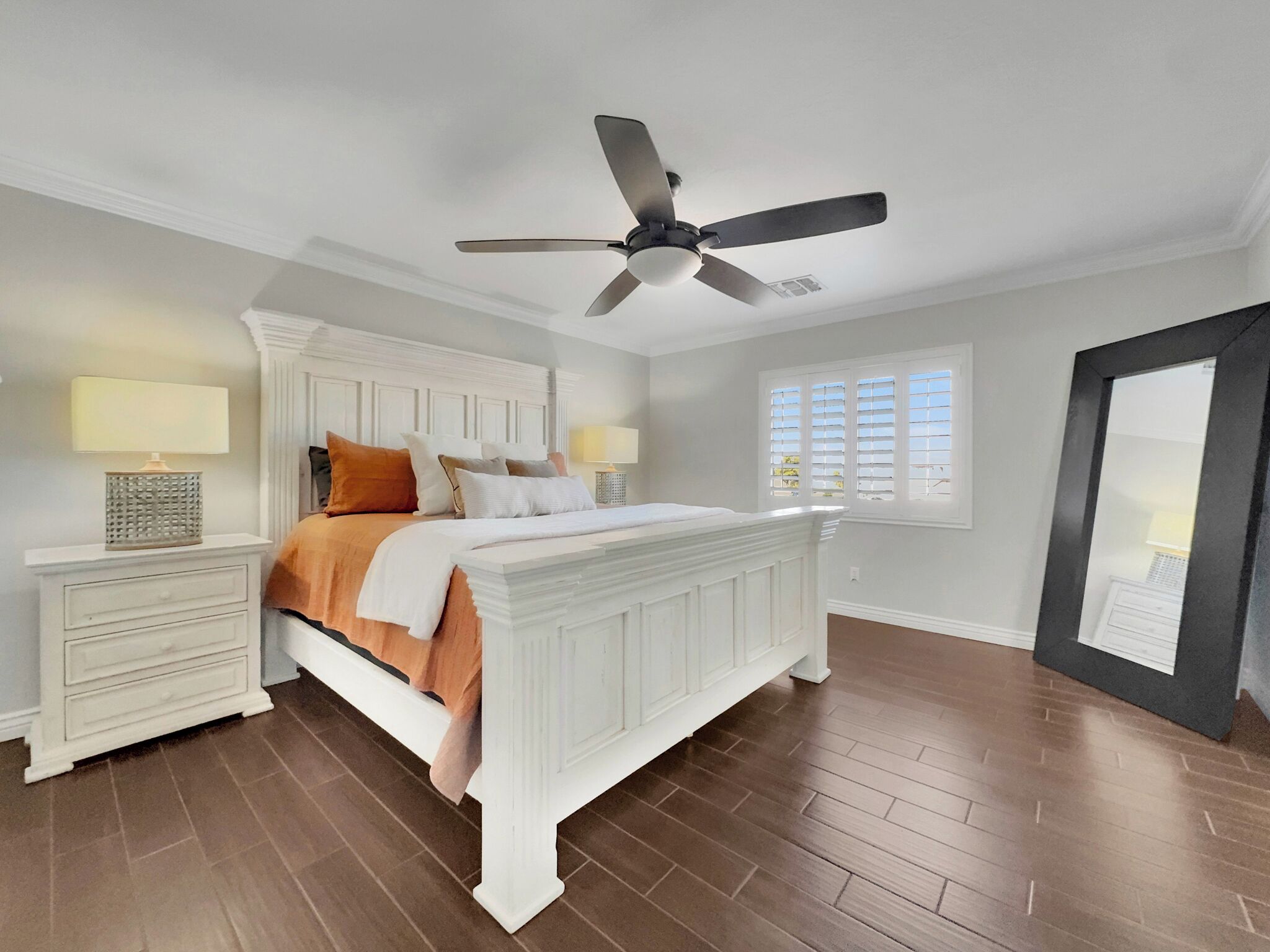 A bedroom with a white bed, two bedside tables, a ceiling fan, a window with blinds, and a door.