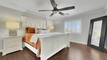 A bedroom with a white bed, two bedside tables, a ceiling fan, a window with blinds, and a door.