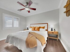 Une chambre à coucher comprenant un lit, des tables de chevet, un ventilateur de plafond et une fenêtre avec des stores.