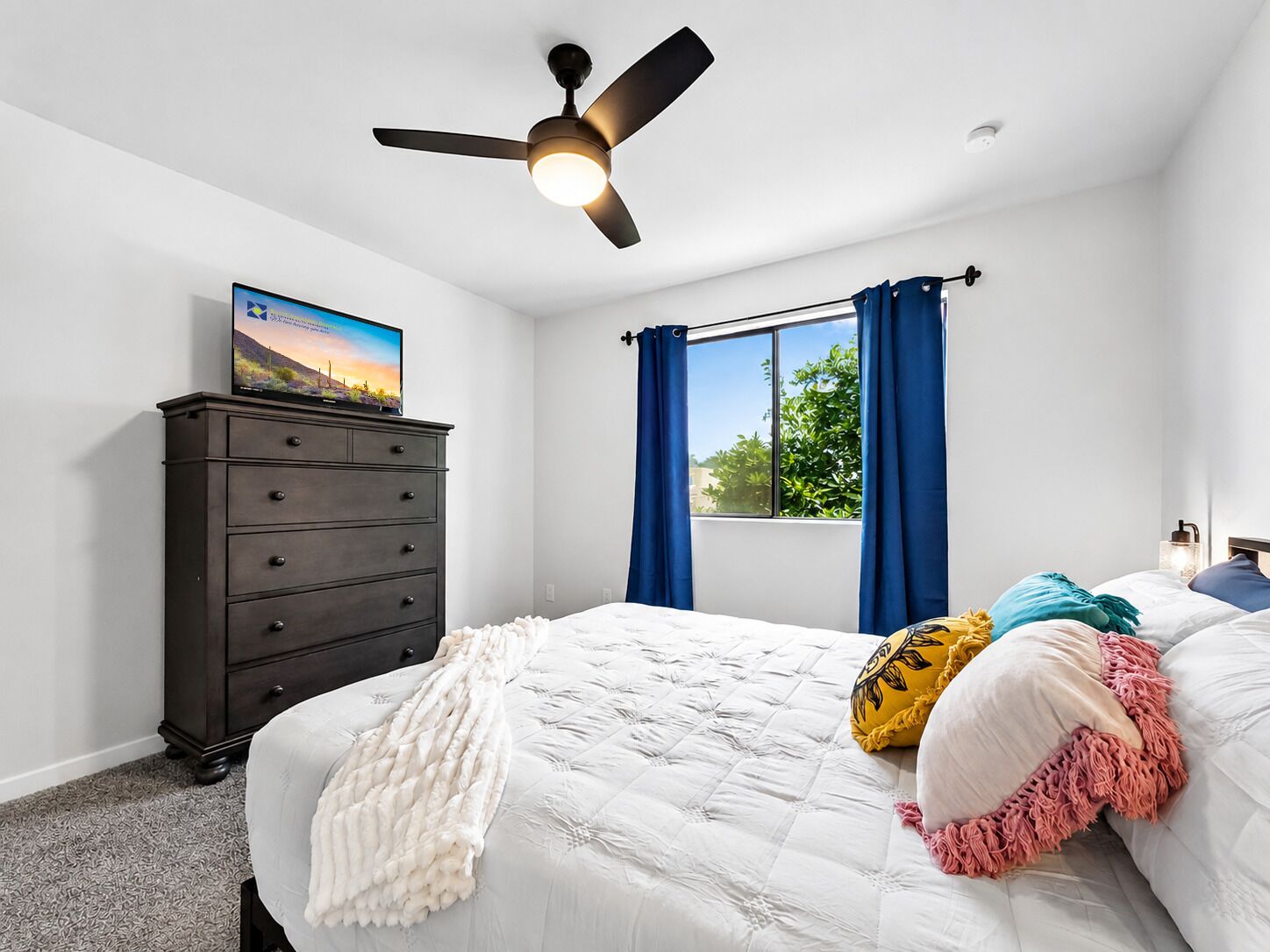 Une chambre à coucher avec un lit, une commode, un ventilateur de plafond, une fenêtre avec des rideaux et un téléviseur posé sur la commode.