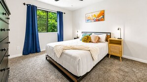 Una camera da letto con un letto, una ventola a soffitto, una finestra con tende, un comodino e un'opera d'arte appesa al muro.