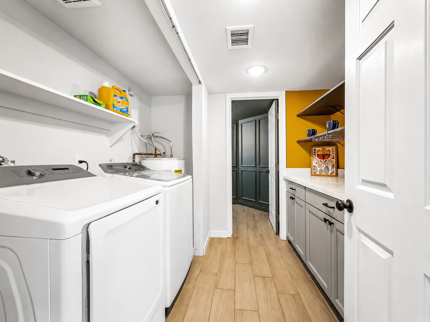 A laundry area with a washing machine, a sink, and a water heater. There is a hallway leading to a kitchen with a yellow accent wall and a door.