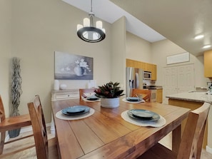 Un comedor con una mesa de madera, sillas, una araña, un cuadro y una cocina al fondo.