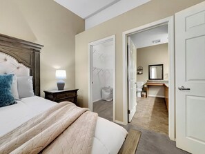 Un dormitorio con cama, mesita de noche, lámpara y una puerta abierta que da al baño.