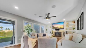 A modern living room with a fireplace, a flat-screen TV, and a large window overlooking a garden. - Yucca Mesa (Mesa)