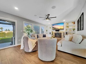 A modern living room with a fireplace, a flat-screen TV, and a large window overlooking a garden. - Yucca Mesa (Mesa)