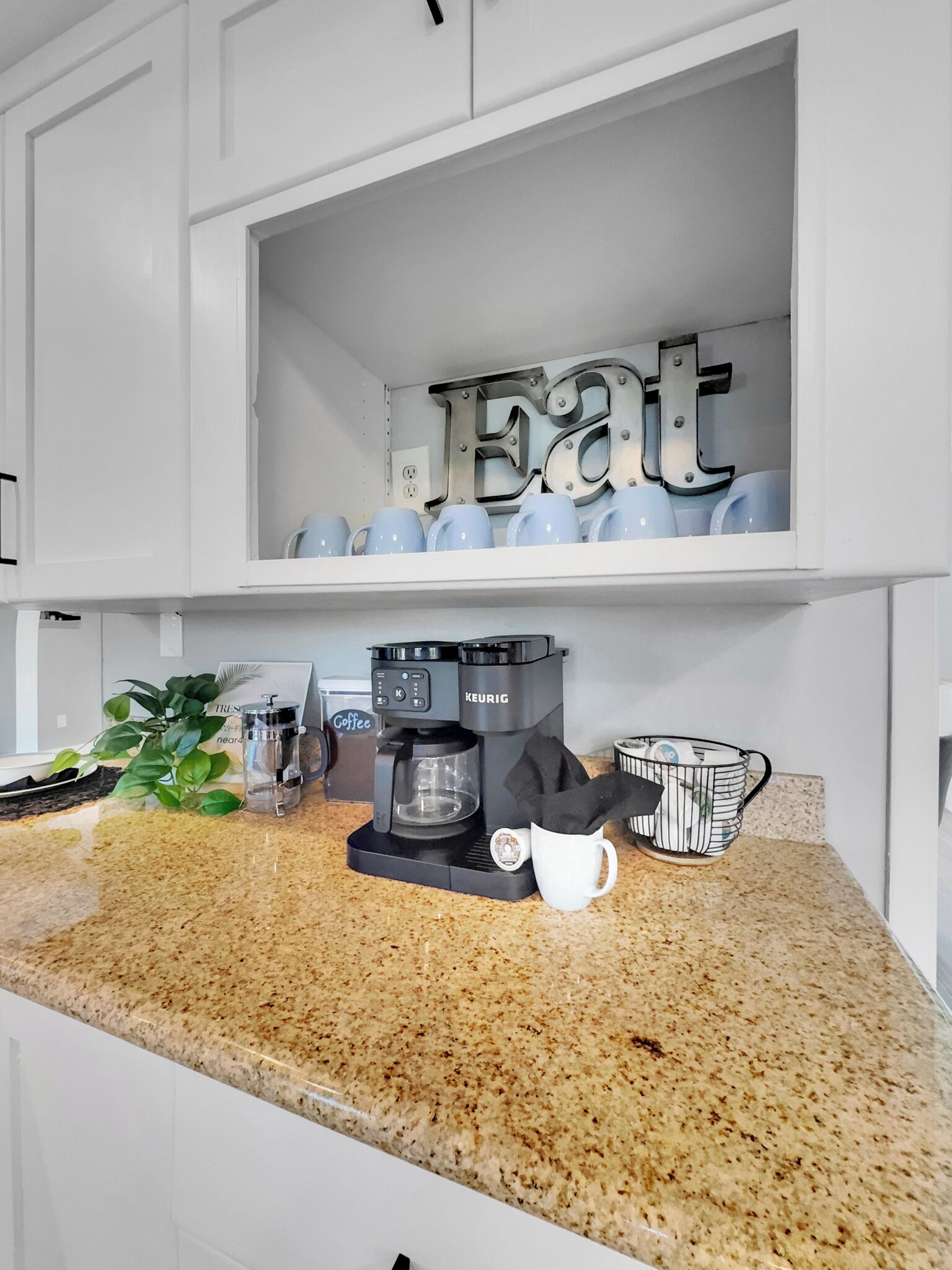 A kitchen with a coffee maker, a plant, and a shelf with cups and a sign that says 'EAT'.