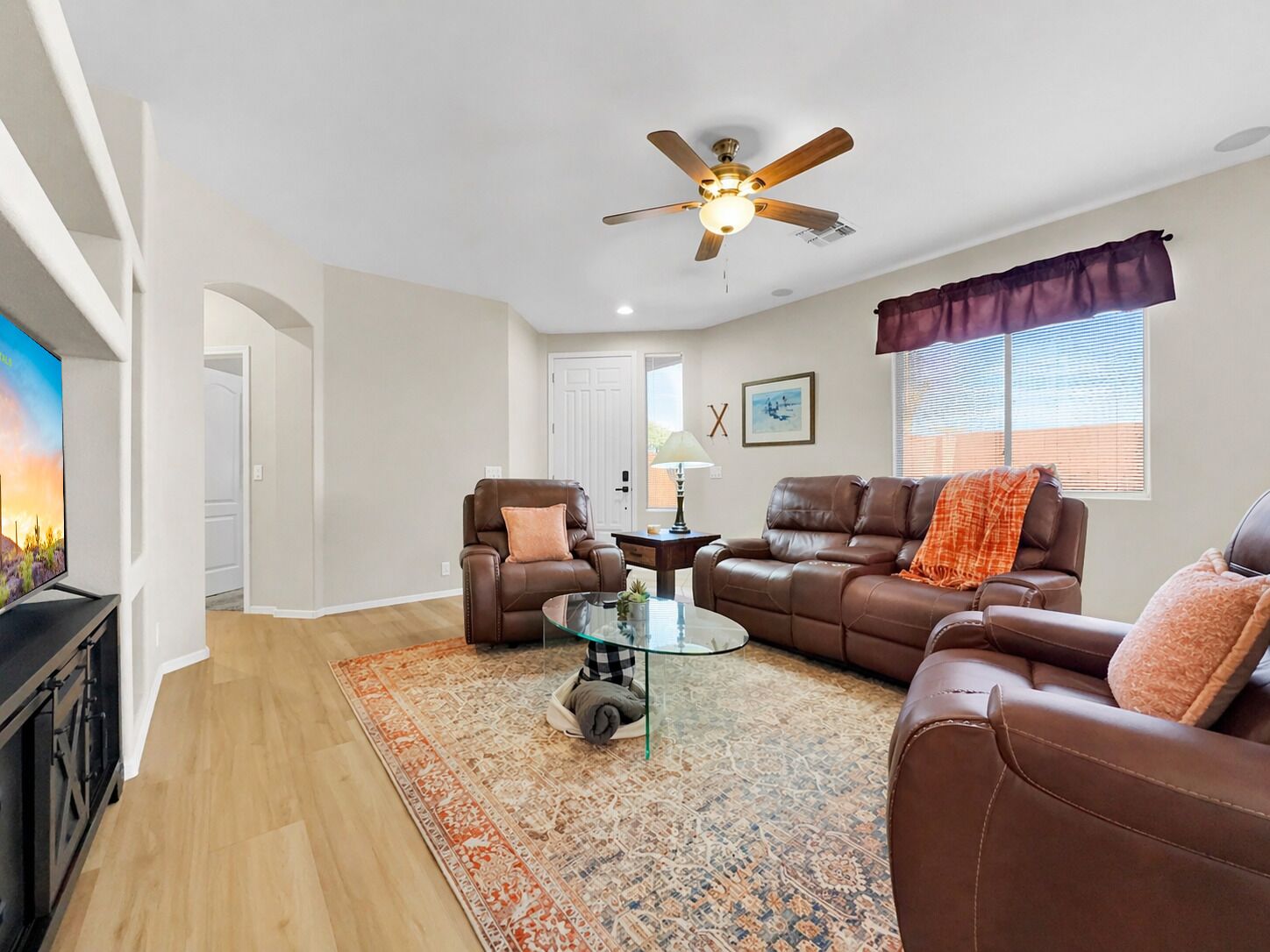 A living room with a ceiling fan, a brown leather sofa, a glass coffee table, and a patterned rug.