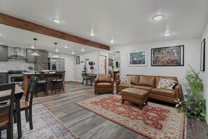 A spacious living area with a brown leather sofa, a dining table with chairs, a kitchen in the background, and a large painting on the wall. - Relaxing Mid-Century Home Near Ski Resorts with SwimSpa & EV Charger (Cottonwood Heights)