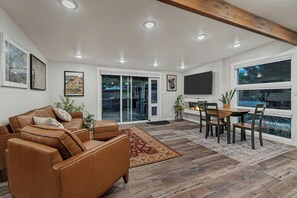 A modern living room with a brown leather sofa, a dining table with chairs, a flat-screen TV, and artwork on the walls. - Relaxing Mid-Century Home Near Ski Resorts with SwimSpa & EV Charger (Cottonwood Heights)