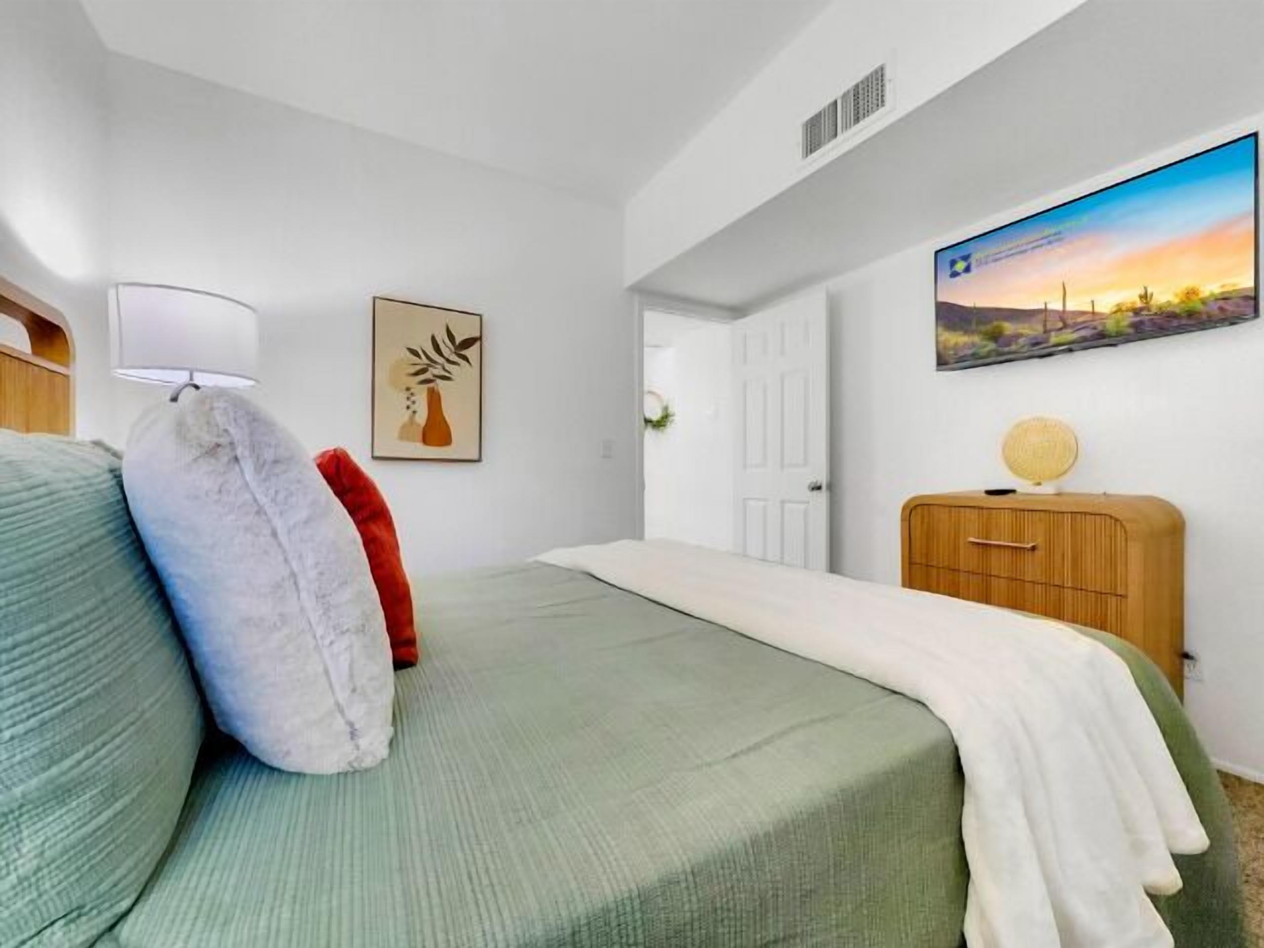 Une chambre à coucher avec un lit, une lampe de chevet, un tableau encadré, une commode et un téléviseur fixé au mur.