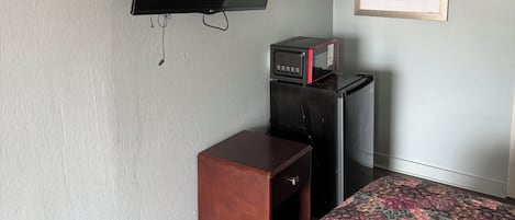 A room with a flat-screen TV mounted on the wall, a microwave on a black cabinet, a small wooden stool, and a bed with a floral bedspread.