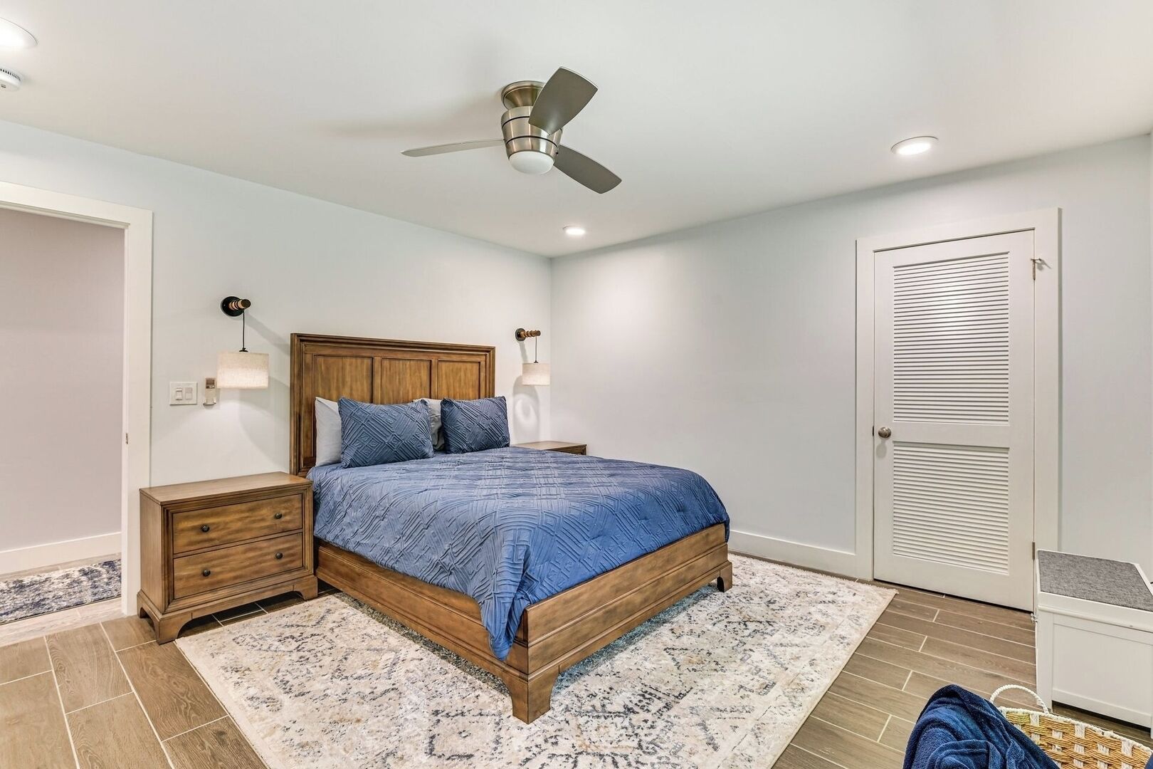 A bedroom with a bed, nightstands, a ceiling fan, a wall-mounted lamp, and a door.