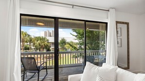 Et værelse med en hvid sofa, en skydedør til en balkon med udsigt til palmer og bygninger.
