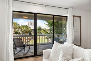 Ein Zimmer mit einem weißen Sofa, einer Schiebeglastür, die auf einen Balkon mit Blick auf Palmen und Gebäude führt.
