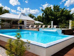 A swimming pool with lounge chairs, a covered seating area, and a building with a sign. - Villa Caporia (Saint François)
