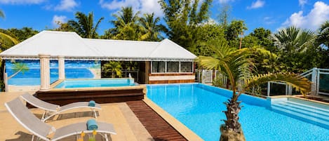 A pool area with lounge chairs, a covered seating area, and a view of the ocean.