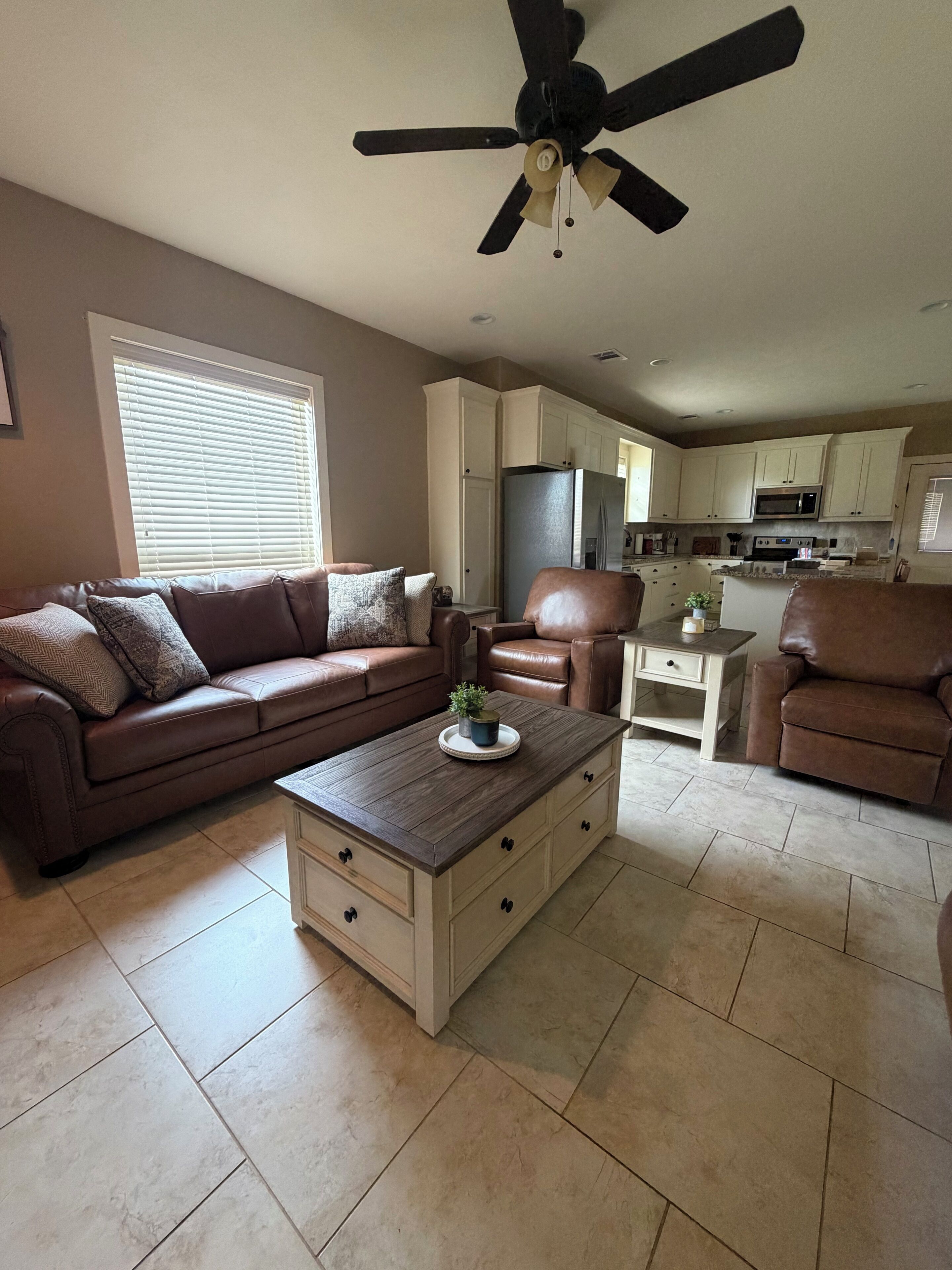 A living room with a brown leather sofa, a coffee table with drawers, a ceiling fan, and a kitchen in the background.