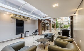 A modern outdoor seating area with a glass ceiling, a TV mounted on the wall, and a wheelchair.