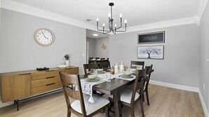 Comedor con mesa de madera dispuesta para cuatro, una lámpara de araña, un reloj y una cita enmarcada en la pared.