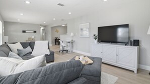 Una sala de estar moderna con un sofá gris, un televisor de pantalla plana sobre un mueble blanco y un comedor al fondo.