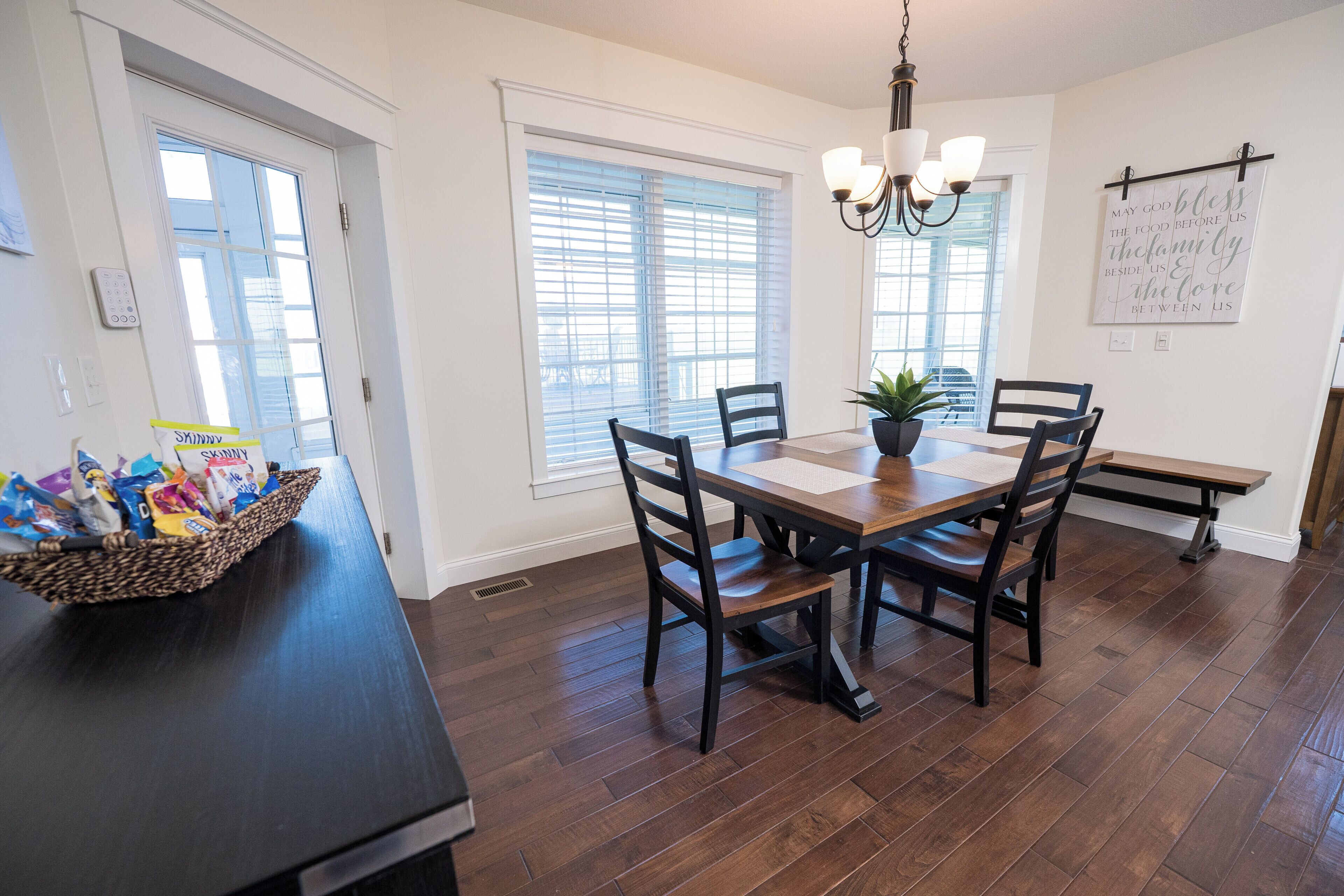 A dining area with a wooden table and chairs, a chandelier, and a basket of snacks on the counter.