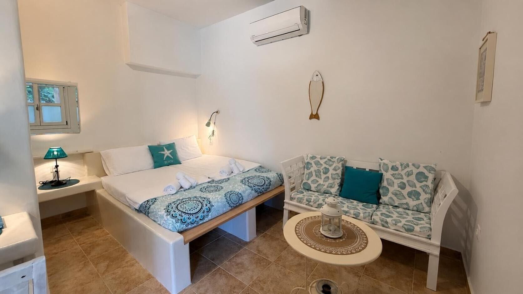 A bedroom with a bed, a nightstand, a lamp, a wall-mounted air conditioner, a sofa, and a small table with a lantern.