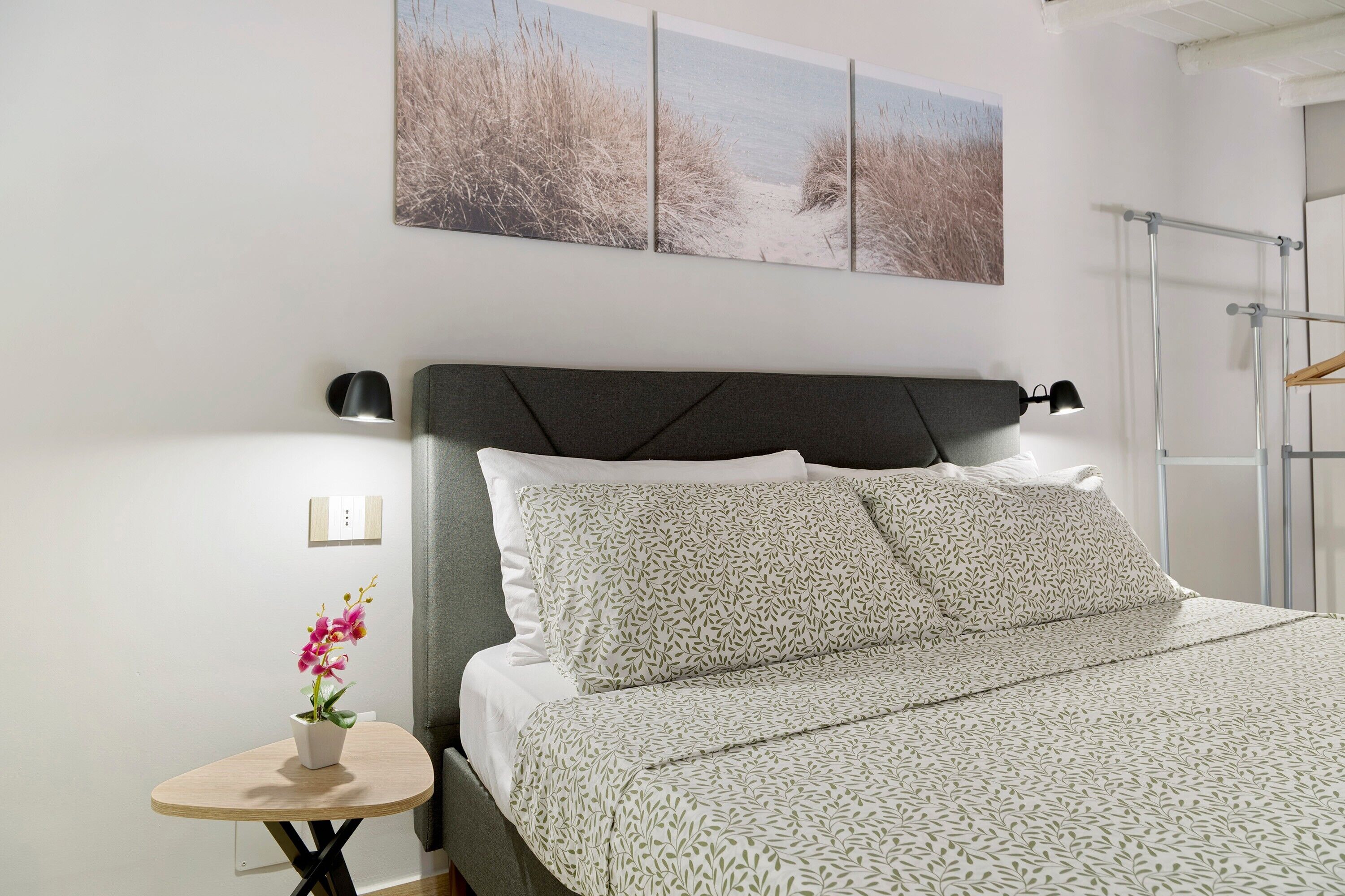 Ein ordentlich bezogenes Bett mit einem gemusterten Überwurf, ein Nachttisch mit einem Blumentopf und eine Wandleuchte.