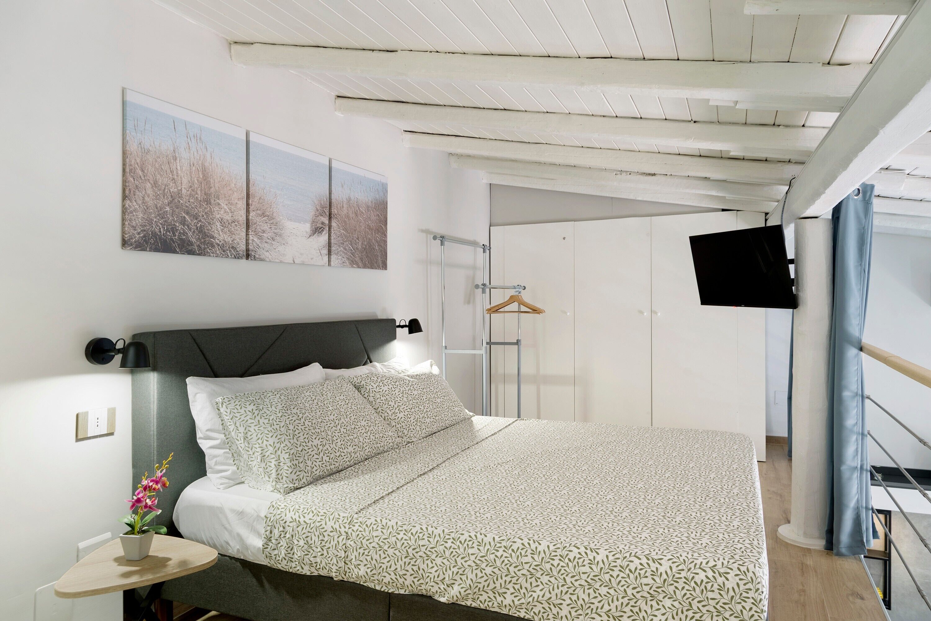 A neatly arranged bedroom with a bed, a bedside table with a lamp, a hanging TV, and a wardrobe.