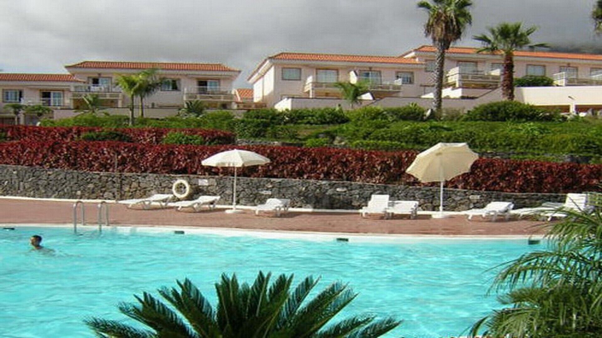 Piscina con sillas de descanso y sombrillas, rodeada de jardines con palmeras y un muro de piedra.