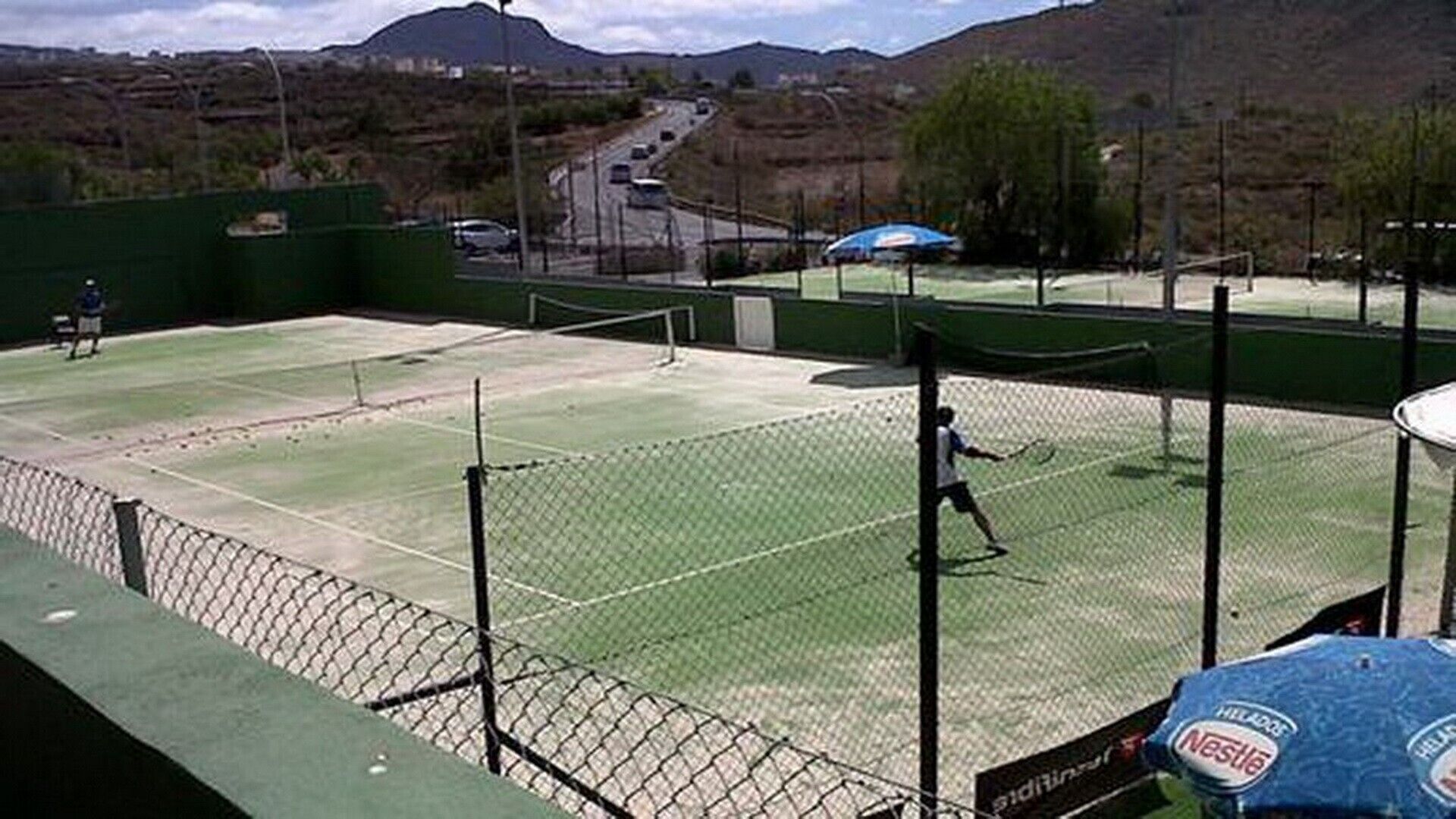 Ein Tennisplatz im Freien mit Netz, umgeben von einem Zaun, und eine Straße im Hintergrund.