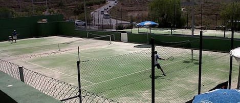 Una cancha de tenis al aire libre con una red, rodeada por una valla, y una carretera al fondo.