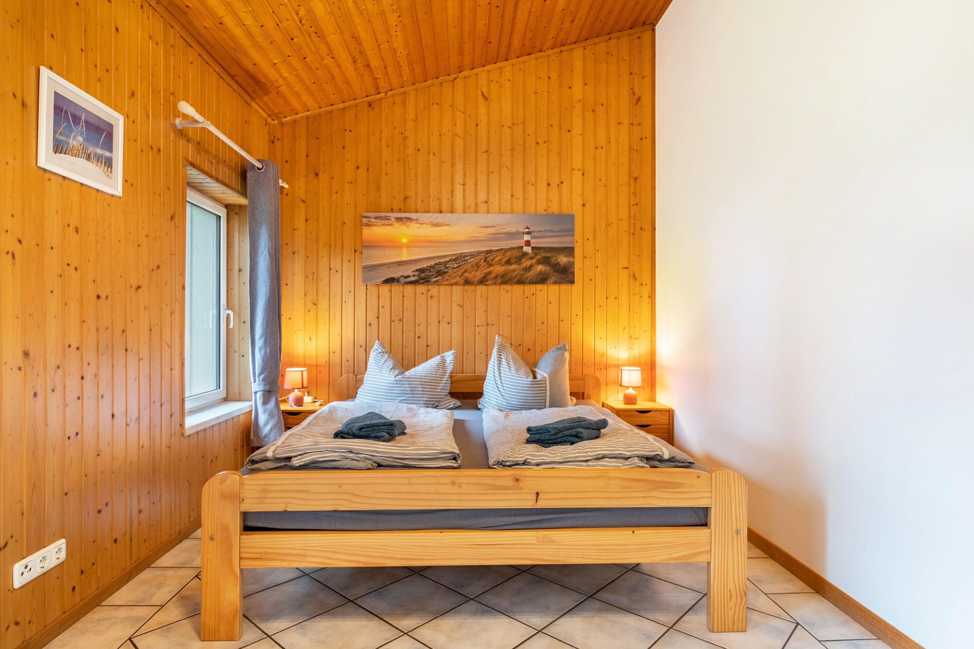 Ein Zimmer mit Holzwänden, einem Bett, zwei Nachttischen, einem Fenster und einem Gemälde, das einen Sonnenuntergang über einem Strand zeigt.
