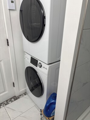 A stacked washer and dryer unit with a control panel, a door, and a blue detergent bottle on a small cart. - 	
Quiet Executive Retreat | Balcony, Parking, Near Hospital & UdeM” (Moncton)