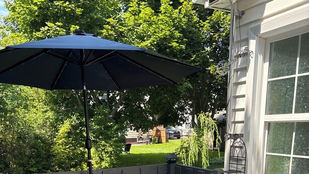 An outdoor seating area with a bench and a table, a large umbrella, and a view of a garden and house. - 	
Quiet Executive Retreat | Balcony, Parking, Near Hospital & UdeM” (Moncton)
