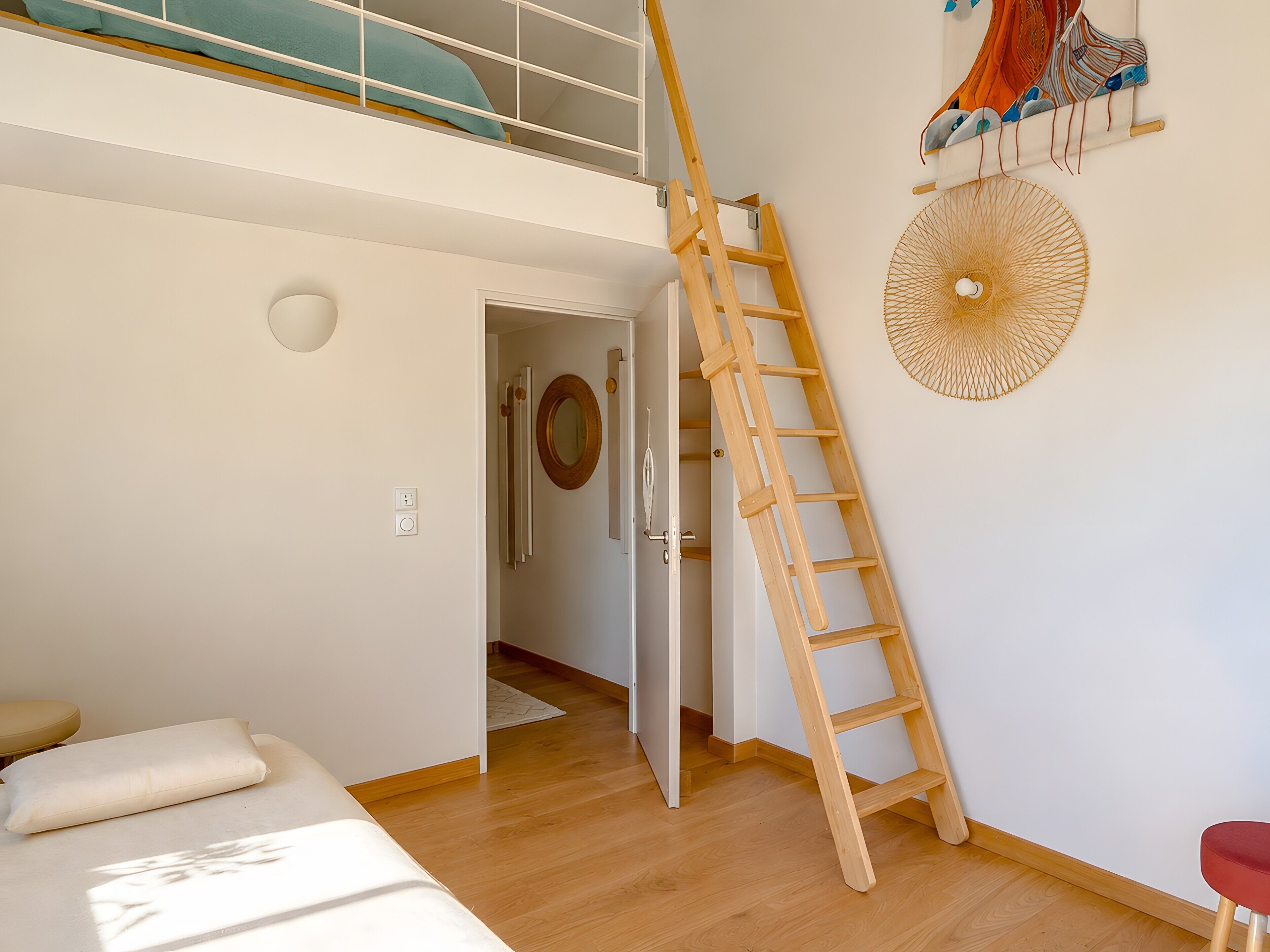 Une pièce avec une échelle en bois, un lit, une porte donnant sur une autre pièce et un ventilateur fixé au mur.