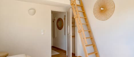 Ein Zimmer mit einer Holzleiter, einem Bett, einer Tür zu einem anderen Raum und einem an der Wand befestigten Ventilator.