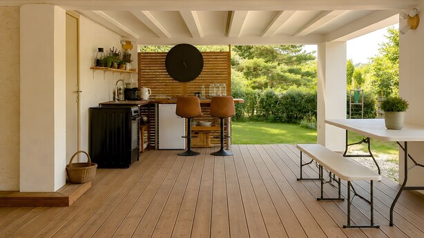 Een terras buiten met een houten terras, een ronde tafel, een bank en een keuken met een spoelbak en kastjes.