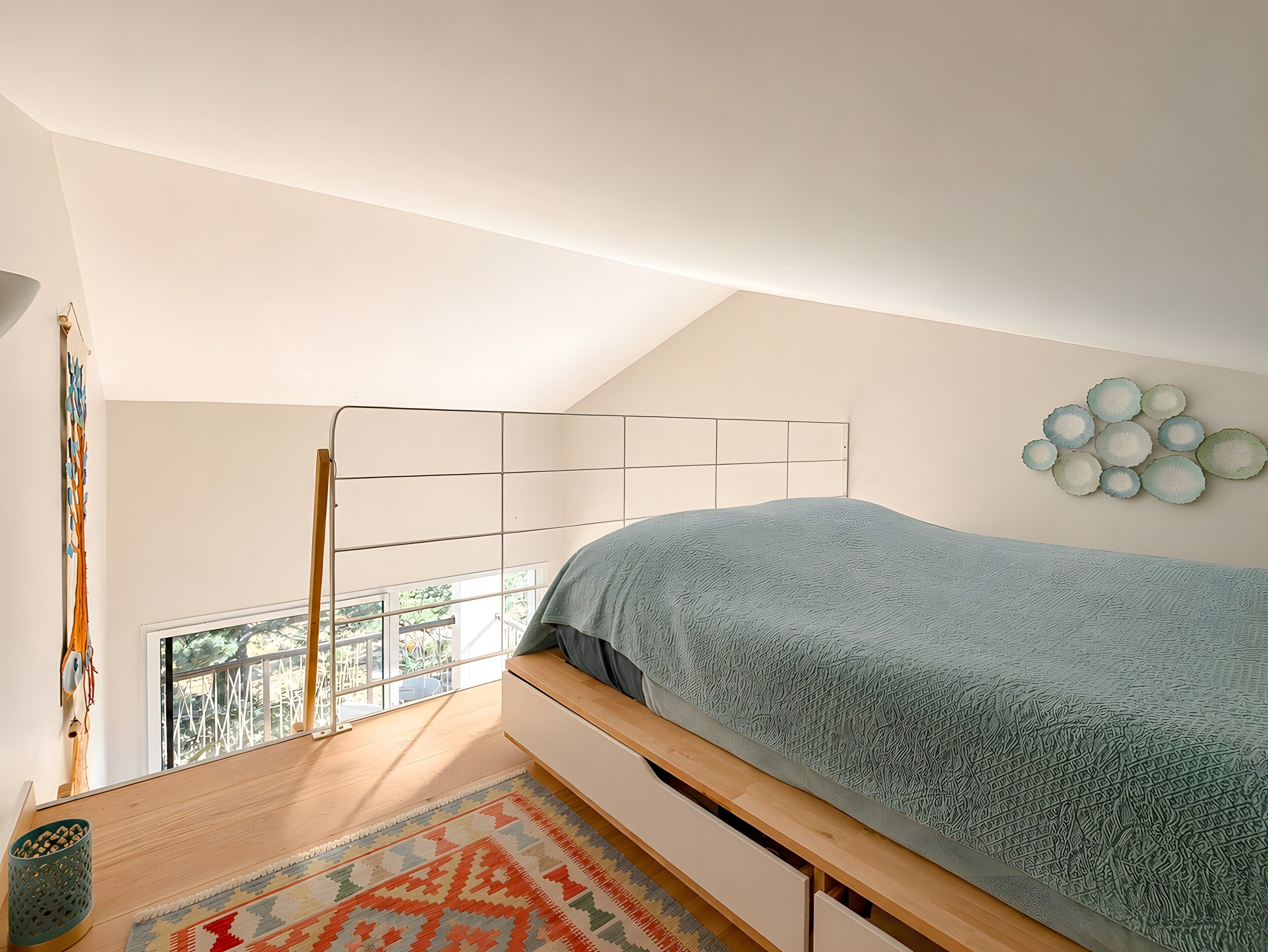 A bedroom with a sloped ceiling, a bed with a patterned bedspread, a wall-mounted light fixture, and a view of the outdoors through a window.