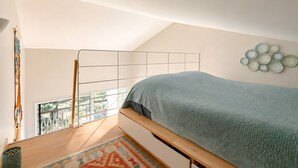 Een slaapkamer met een schuine kap, een bed met een decoratieve dekbedovertrek, een wandlamp en uitzicht op de omgeving door een raam.