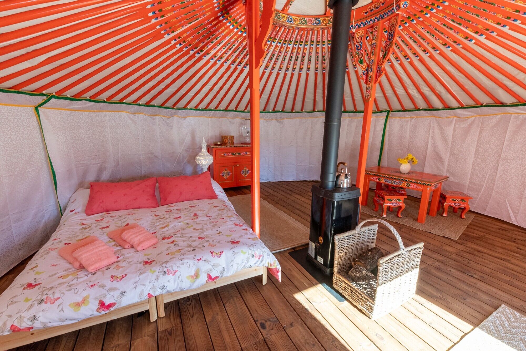 Ein gemütliches Zimmer mit einem Bett, einem Tisch, einem Stuhl, einem Korb und einem Kaminofen.