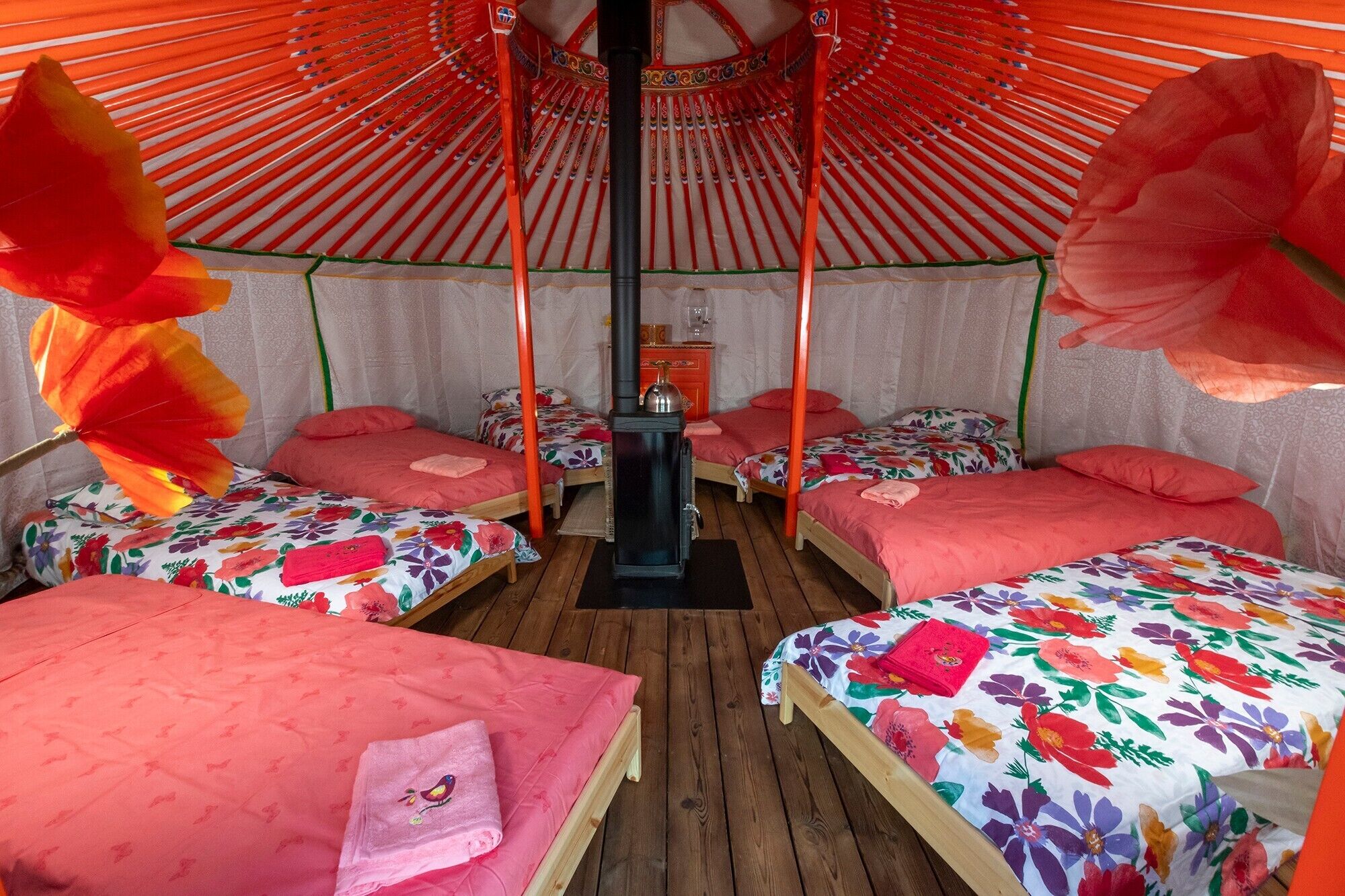 Une chambre avec quatre lits, chacun recouvert d’une couvre-lit à motifs floraux et agrémenté d’un oreiller rose. On y trouve une table en bois placée au centre et un radiateur noir.