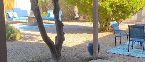 A covered patio with wicker furniture, a ceiling fan, and a view of a garden with trees and a blue sofa.