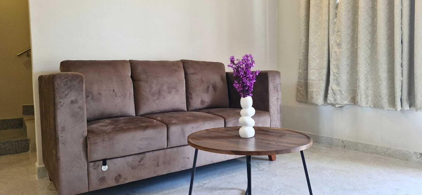 Un sofá marrón con un jarrón blanco lleno de flores moradas sobre una mesa redonda de madera.