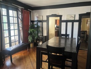 Un comedor con una mesa y sillas de madera, un banco, una planta en maceta y vistas a través de una ventana con cortinas.