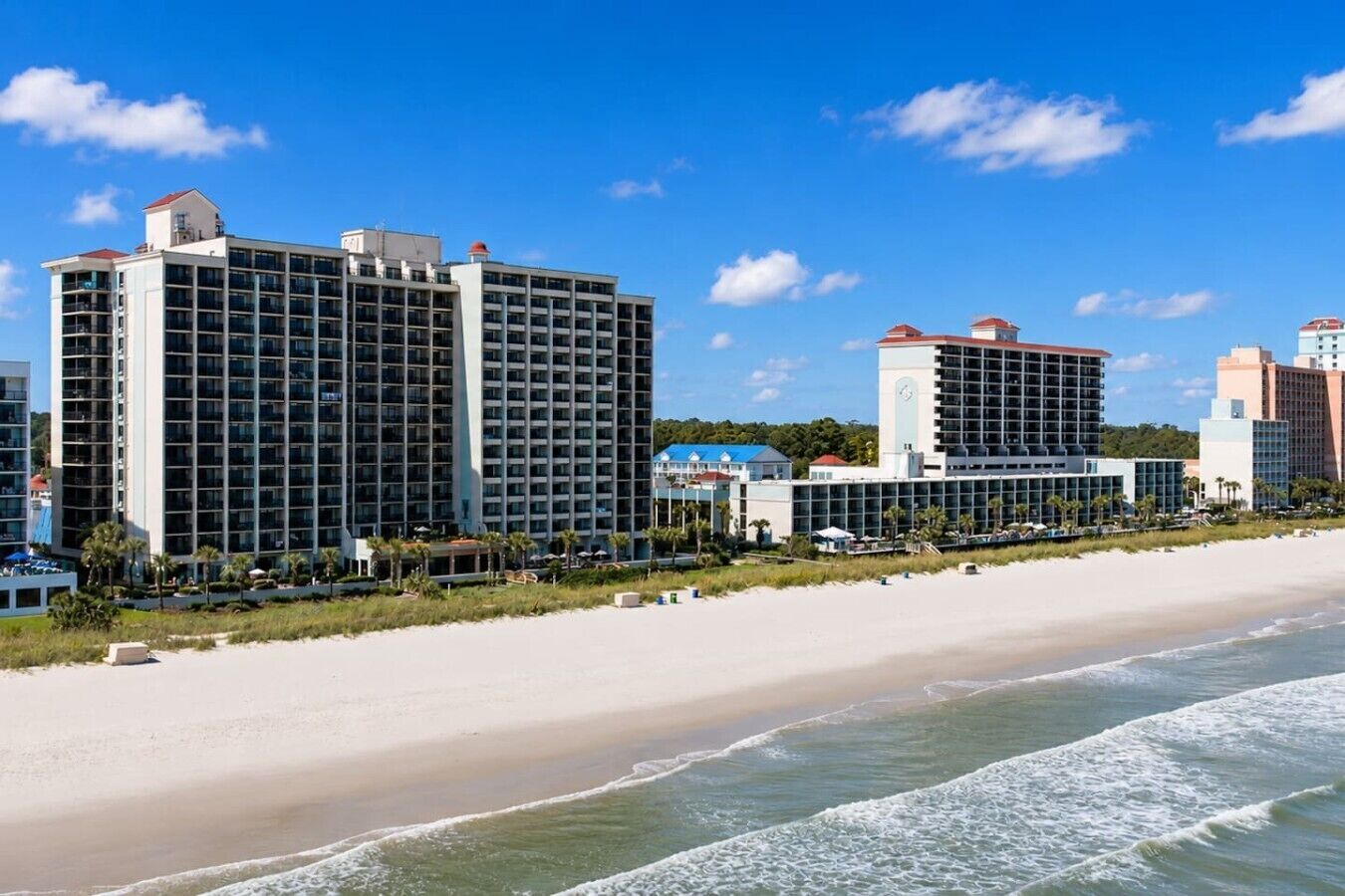 Et strandhotel med flere høje bygninger, en sandstrand og havnebølger.