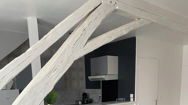 Um espaço de estar moderno com um sofá cinzento, uma mesa de centro de madeira e uma cozinha com bancada branca e eletrodoméstico preto.