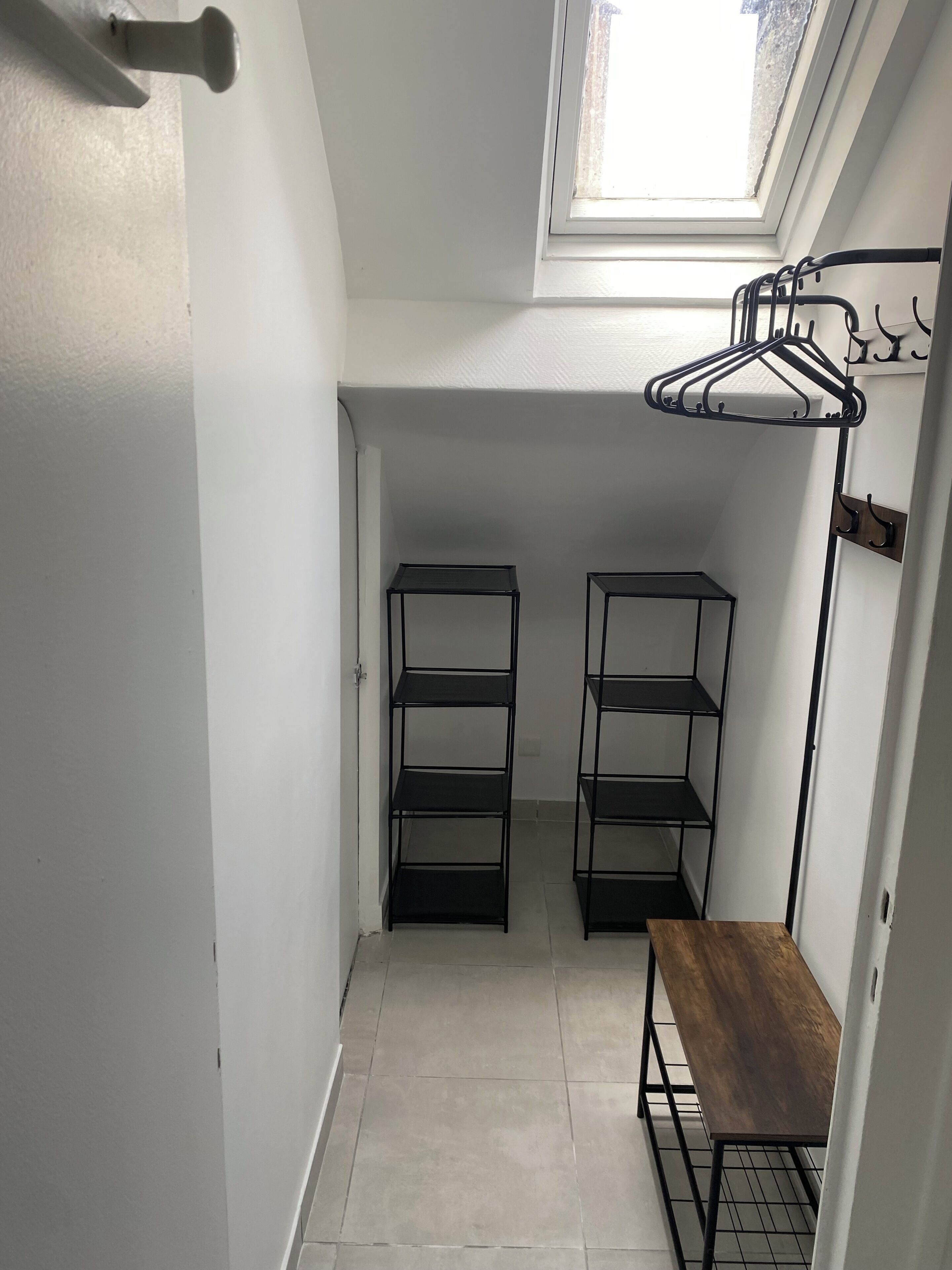 A compact room with a skylight, two black metal shelving units, a wooden bench, and a clothing rack.