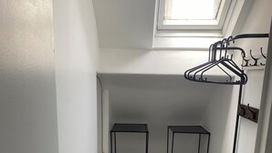 Quarto compacto com claraboia, duas estantes de metal preto, um banco de madeira e um cabideiro.